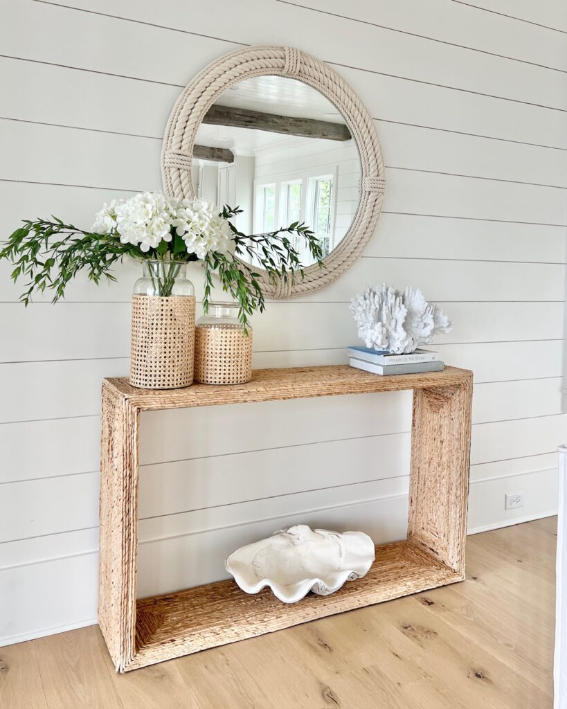styled modern coastal console table with a round rope mirror, woven vases with white hydrangeas, a coral sculpture, and a giant clamshell accent