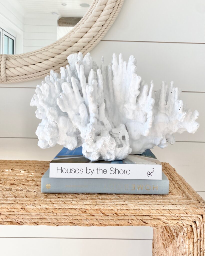 Coastal-inspired decor featuring a white coral sculpture stacked on coffee table books, styled on a woven console table. A perfect blend of natural textures for a modern coastal home.